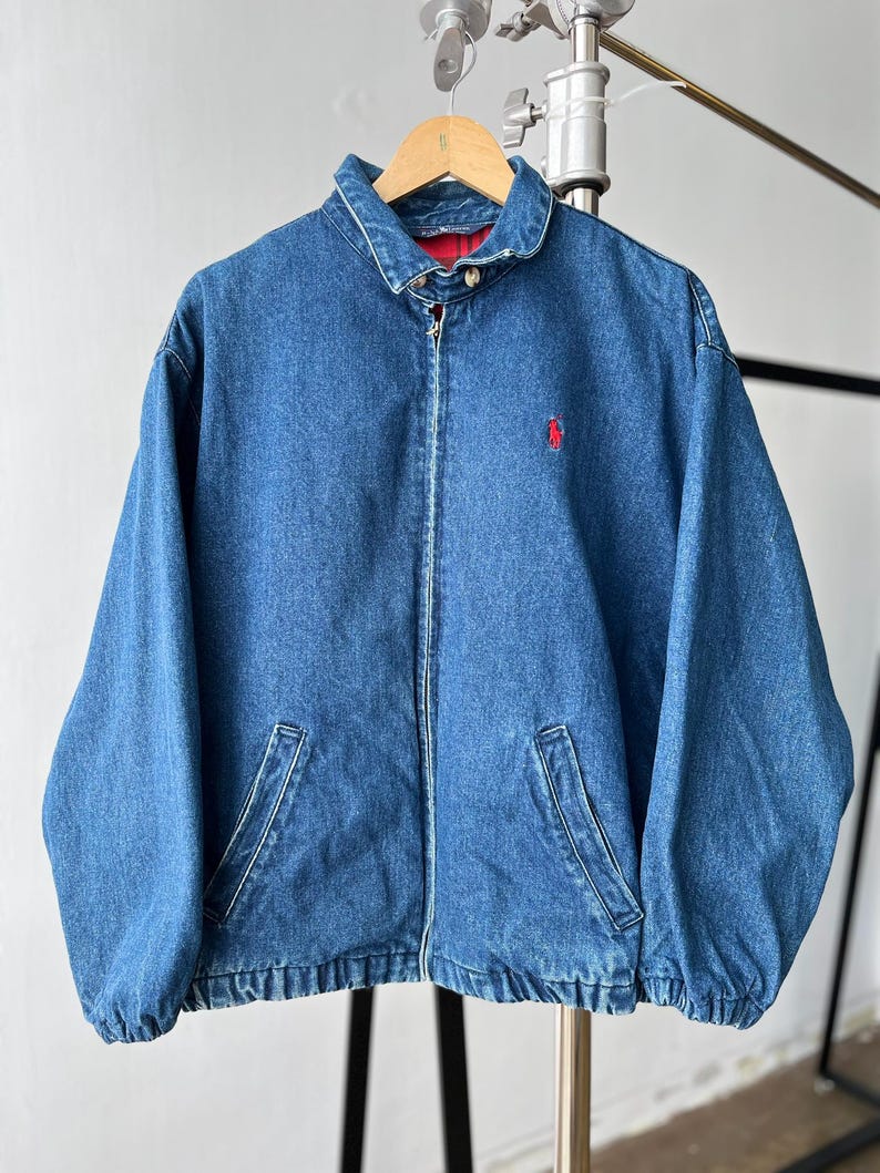 May include: A blue denim jacket featuring a full-length front zipper, a fold-down collar, and two slanted front pockets. The jacket has a red embroidered logo on the left chest and a red and black plaid lining visible at the inner collar area.