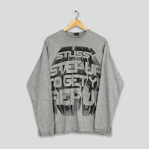 Peut inclure: Un t-shirt gris à manches longues avec un motif graphique. Le motif présente le mot "STUSSY" en grandes lettres en gras, ainsi que les mots "STEP UP TO GET REP". Le t-shirt est suspendu à un cintre en bois.