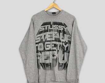 May include: A gray long-sleeve shirt with a graphic design. The design features the word "STUSSY" in large, bold letters, along with the words "STEP UP TO GET REP". The shirt is hanging on a wooden hanger.