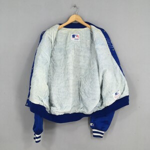 May include: A royal blue Starter brand varsity jacket with a white quilted interior lining. The jacket features a snap-button front closure, long sleeves with striped ribbed cuffs, and a Major League Baseball logo patch on the inner collar.