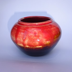 May include: A red and brown ceramic pot with a rounded shape and a slightly flared rim. The pot has a mottled glaze with shades of red, brown, and white.