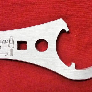 Könnte beinhalten: Ein silberner Schraubenschlüssel mit drei verschiedenen Öffnungen. Der Schraubenschlüssel trägt den Text "PUSH PERPENDICULAR TO WRENCH" und einen Pfeil, der nach rechts zeigt.