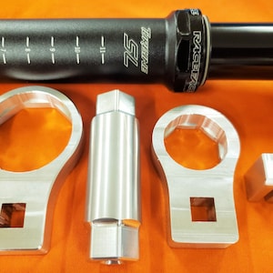 Puede incluir: Un juego de cuatro herramientas de color plateado para ajustar una horquilla de suspensión de bicicleta. Las herramientas incluyen una llave, un casquillo, una llave hexagonal y un espaciador. Las herramientas están sobre una superficie de color naranja brillante.