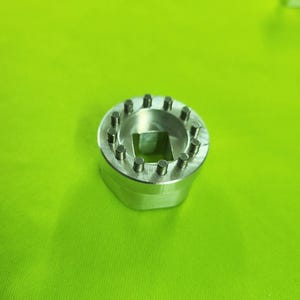 May include: A silver metal tool with a circular top featuring evenly spaced cylindrical studs. The center has a square opening. The tool is set against a bright green background.