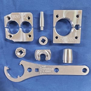 以下が含まれることがあります： 「2026 X2」の文字が入ったクランプのような部品2つ、説明付きのレンチ、その他いくつかの幾何学的な部品など、光沢のある銀色の金属製機械部品の詰め合わせ。部品は青い表面に配置されています。