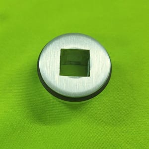 Puede incluir: Un casquillo metálico plateado con una abertura cuadrada en el centro. El casquillo tiene forma circular con un anillo de goma negro alrededor del borde. El fondo es de color verde liso.