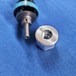 Puede incluir: Primer plano de un componente mecánico desmontado. La parte superior presenta una pieza cilíndrica negra y turquesa. Debajo, una varilla roscada metálica se extiende hacia abajo. Una tuerca hexagonal plateada está colocada a la derecha.