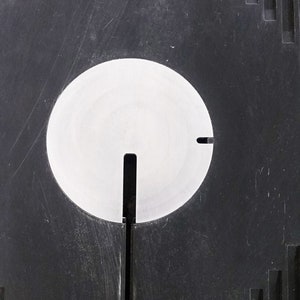 May include: A black plastic surface with a circular cutout. The cutout is filled with a white, round, flat disc with a vertical slot in the center.