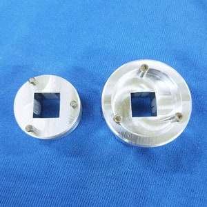 Puede incluir: Dos punzones cuadrados de metal plateado con cuatro puntas en el borde exterior. El punzón más grande tiene un diámetro de unos 5 cm y el más pequeño de unos 3,8 cm.