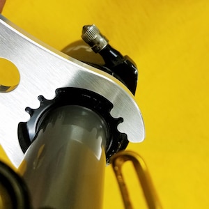 May include: A silver metal bicycle tool with various shaped openings is shown against a bright yellow background. The tool is positioned over a black component, possibly a part of the bicycle's valve stem.