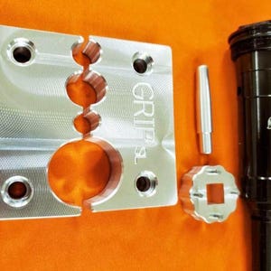 Puede incluir: Una herramienta de metal plateado con un agujero cuadrado y un agujero circular, una varilla de metal plateado, una herramienta hexagonal de metal plateado y una herramienta de metal negro con el texto "FOX GRIPS" en ella. La herramienta de metal plateado tiene el texto "GRIP SL" en ella.