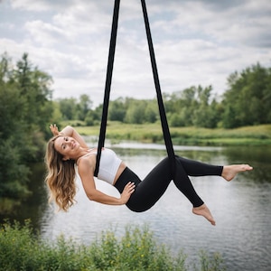 Puede incluir: Un top deportivo corto de color blanco y unos leggings negros de talle alto, usados mientras se está suspendido en una hamaca de yoga aéreo negra. La tela es de color sólido con una textura suave, diseñada para flexibilidad y soporte.
