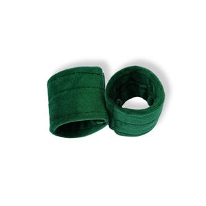 May include: A pair of dark green wristbands made of a soft, plush fabric. Each circular band features a thick, padded construction with visible horizontal stitching lines running along the exterior surface, creating a textured, cylindrical appearance.