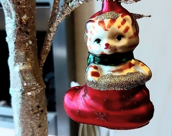 May include: A red and white glass Christmas bauble shaped like a cat wearing a green scarf and sitting in a red stocking. The bauble is hanging from a gold glitter branch.