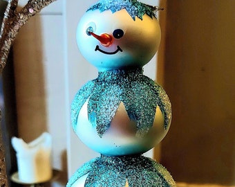Pode incluir: Um enfeite de vidro de boneco de neve de três peças com acabamento em glitter azul e branco. O boneco de neve tem um nariz vermelho e um rosto sorridente.