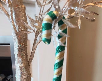 Pode incluir: Um enfeite de bengala de açúcar com glitter verde e dourado pendurado em um galho de glitter prata.