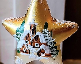 Pode incluir: Um enfeite de Natal em forma de estrela dourada com uma cena pintada de uma casa coberta de neve com um campanário de igreja. O enfeite é coberto com glitter branco.