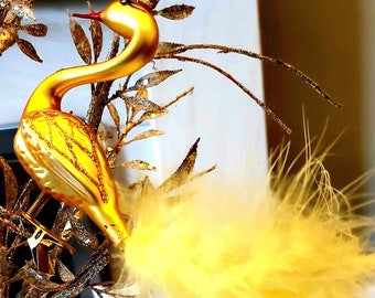May include: A gold-coloured bird ornament with a long neck and a yellow feathery tail. The bird is perched on a gold-coloured branch with leaves. The ornament is likely for holiday or home decor.
