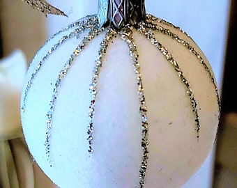 May include: A white Christmas bauble with silver glitter stripes. The bauble is round and has a silver metal top.