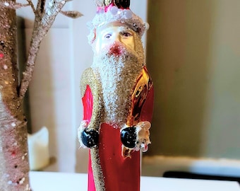 Pode incluir: Um enfeite de Natal vintage de vidro que representa o Papai Noel com uma túnica vermelha com detalhes dourados e uma barba branca. Ele está segurando um cajado em uma mão e uma bolsa na outra.