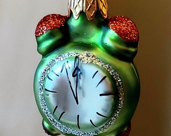 May include: Green glass Christmas bauble shaped like an alarm clock with a white face and black hands. The clock face has a silver glitter border. The bauble has a gold top and red glitter accents.