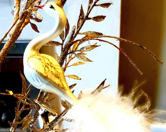 May include: A white glass bird ornament with gold accents and a white feather tail. The bird is perched on a gold twig with a gold glitter finish.