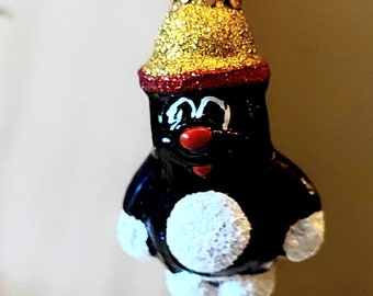 May include: A black and white Christmas bauble shaped like a cartoon character wearing a gold glitter hat with a red band. The character has a white tummy and a red nose.