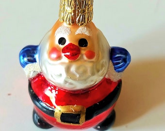 May include: A glass Christmas bauble shaped like Santa Claus. He is wearing a red suit with a black belt, white beard, and a gold hat. He has a red nose and blue eyes.