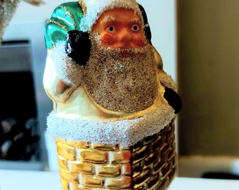 May include: A glass Christmas bauble featuring Santa Claus in a gold brick chimney. Santa is wearing a white hat, a green and black coat, and a white beard. The bauble is covered in glitter.