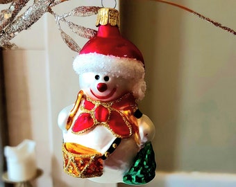 Pode incluir: Um enfeite de Natal de boneco de neve branco com um chapéu vermelho, um cachecol vermelho e dourado e um tambor dourado. O boneco de neve está segurando uma bolsa verde em uma mão.