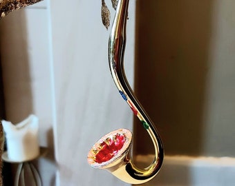 Pode incluir: Um enfeite de Natal de vidro dourado em forma de trombeta com uma campainha vermelha e branca. O enfeite tem um acabamento brilhante.