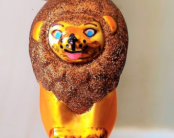 Pode incluir: Um enfeite de Natal de vidro dourado em forma de leão com olhos azuis e língua rosa. O enfeite é coberto de glitter.