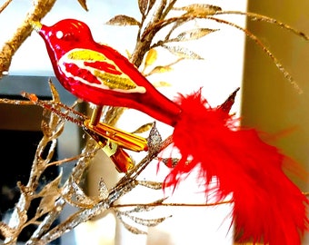 May include: A red glass bird ornament with gold accents and a red feather tail. The bird is perched on a gold twig.