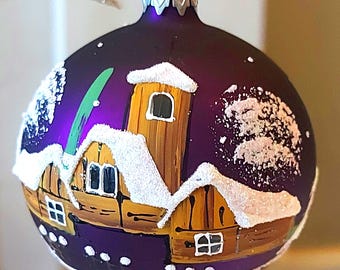 May include: A round, purple Christmas bauble with a hand-painted winter village scene. The village features wooden buildings with snow-covered roofs and a green accent. The bauble has a silver metal cap and a loop for hanging.