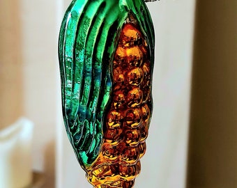 Pode incluir: Um enfeite de Natal de vidro verde e dourado em forma de espiga de milho. O enfeite tem uma parte superior dourada e está pendurado em um galho prateado.