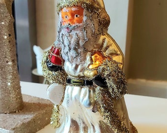 Pode incluir: Um enfeite de Natal de vidro vintage que representa o Papai Noel com uma túnica branca com detalhes dourados, segurando um presente vermelho e dourado. O enfeite tem uma barba branca fosca e um rosto vermelho.