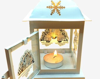 Pode incluir: Uma lanterna de metal branco com um recorte de floco de neve e uma pequena porta. No interior, uma vela de chá acesa emite um brilho quente. A lanterna apresenta uma cena de inverno decorativa nos painéis de vidro.