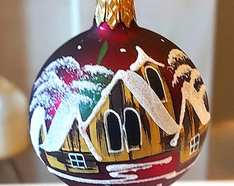 May include: A round Christmas bauble with a red background and a painted winter scene. The scene depicts a snow-covered village with yellow and brown buildings. The bauble has a gold cap and is suspended by a gold wire.