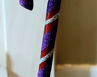 May include: A festive candy cane decoration with a glossy red finish, adorned with purple glitter and silver stripes. This Christmas ornament is a classic holiday symbol, ideal for adding a touch of seasonal cheer.