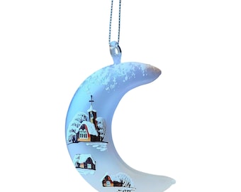Pode incluir: Enfeite de Natal em forma de meia-lua. O enfeite azul claro apresenta uma cena de vila de inverno com uma igreja e casas, todas cobertas de neve. Ele pende de um cordão prateado.