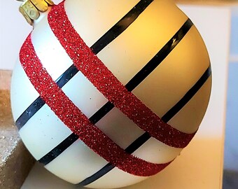 Pode incluir: Enfeite de Natal creme com uma tampa dourada e um laço para pendurar. O enfeite apresenta um padrão cruzado de listras pretas e listras de glitter vermelho brilhante, criando um design festivo.