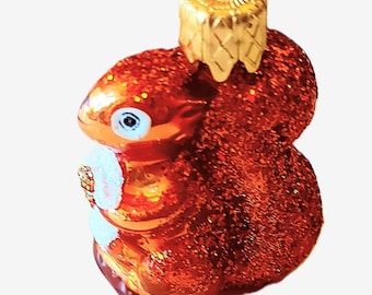 May include: A Christmas tree ornament in the shape of a squirrel. The ornament is predominantly orange with glitter, a white belly, and a gold cap. The squirrel is holding a small, white object. Designed to be hung from a tree.