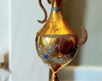 May include: Gold glass Christmas bauble shaped like a vase with a handle and a star design. The bauble is covered in glitter.