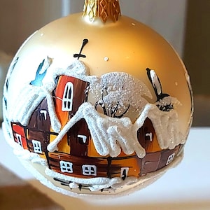 May include: A round, gold-colored Christmas ornament with a winter village scene. The houses have snow-covered roofs and are painted in shades of brown, orange, and red. A small bird is painted on the side. The ornament has a gold cap.
