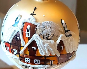 Pode incluir: Enfeite de Natal dourado com uma cena de vila de inverno. As casas, pintadas em tons de castanho, laranja e vermelho, têm telhados cobertos de neve. Um pequeno pássaro é retratado. O enfeite tem uma tampa dourada.