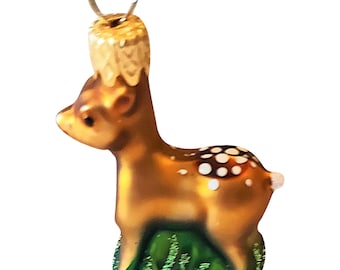 May include: A glass Christmas ornament in the shape of a fawn. The fawn is brown with white spots and stands on green glittery grass. It has a gold cap and a silver loop for hanging.
