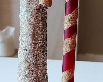 Pode incluir: Enfeite de bengala de doces vermelha e dourada com listras brilhantes. A bengala de doces é uma decoração de Natal clássica, com um topo curvo e uma parte inferior reta. O enfeite está ao lado de uma árvore brilhante.