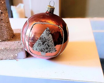 Pode incluir: Um enfeite de Natal redondo, de cor cobre, com um design de árvore de Natal branco e brilhante.