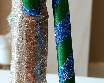 May include: A green candy cane with blue glitter stripes. The candy cane is hanging from a branch of a glittery tree.