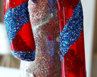 Pode incluir: Um enfeite de bengala de caramelo vermelho e azul com glitter. A bengala de caramelo é decorada com listras de glitter azul.